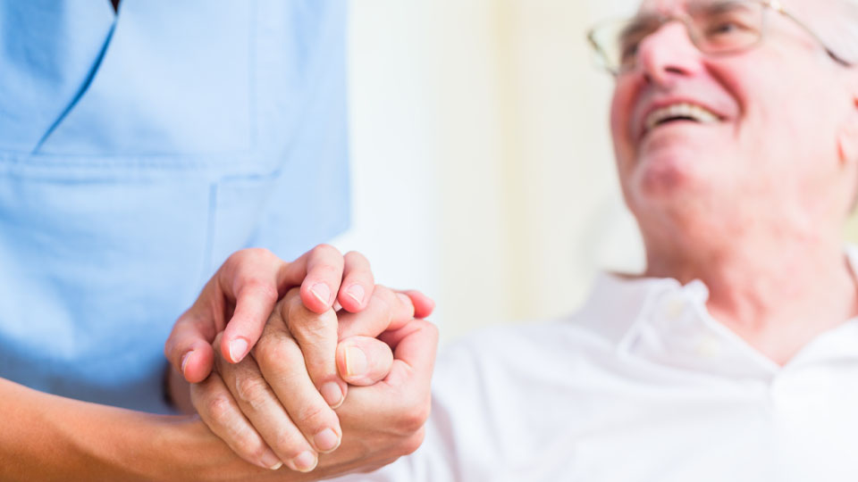 Eine Pflegekraft in blauer Kleidung hält die Hand eines älteren Mannes, der sie anlächelt