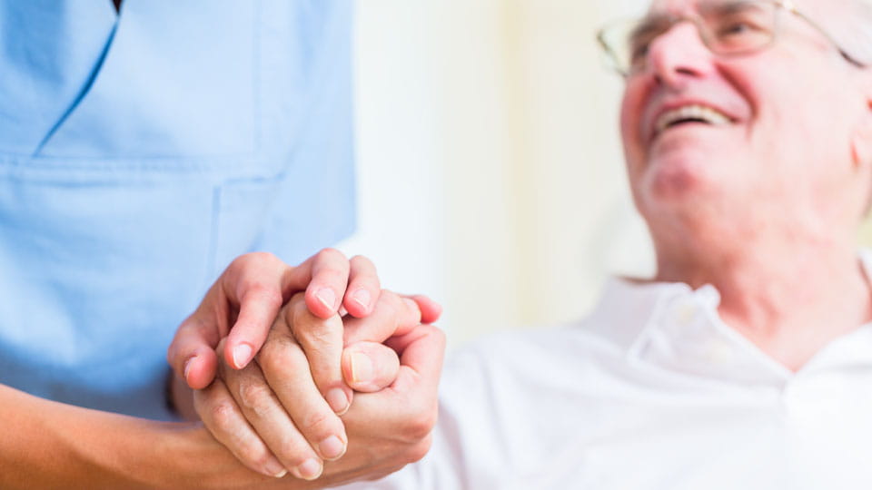 Eine Pflegekraft in blauer Kleidung hält die Hand eines älteren Mannes, der sie anlächelt