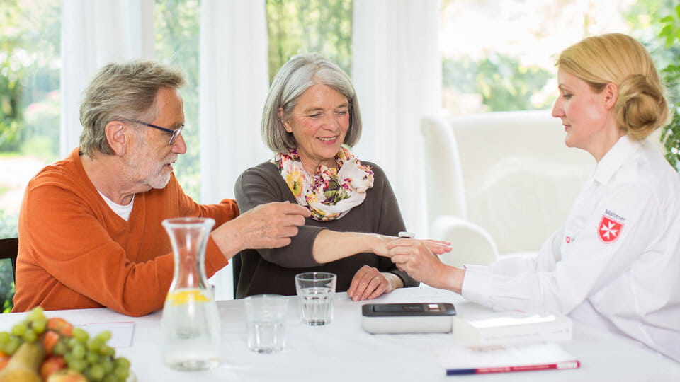 Älteres Paar sitzt am Tisch und spricht mit einer Malteser-Mitarbeiterin, die der Frau die Hand hält