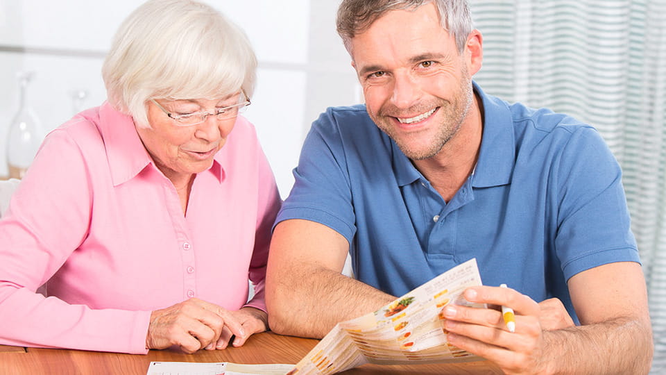 Seniorin und Mann sitzen lächelnd am Tisch und schauen gemeinsam in einen Speiseplan