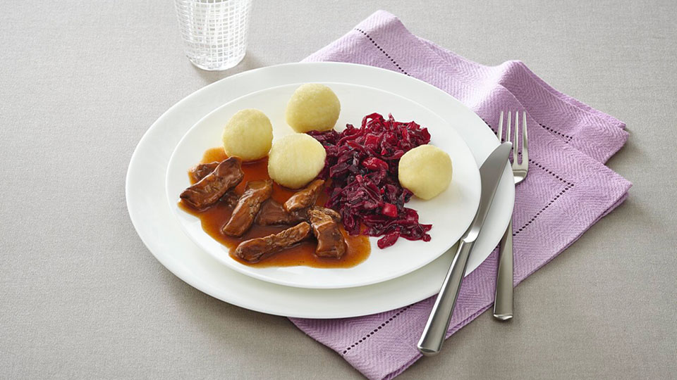 Gericht mit Fleisch in brauner Soße, Kartoffelklößen und Rotkohl auf einem lila Tischset